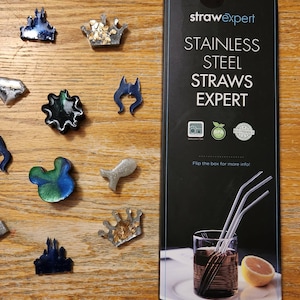 May include: A box of stainless steel straws with the text "strawexpert STAINLESS STEEL STRAWS EXPERT" on the front. The box also has three logos: "Dishwasher Safe", "BPA Free", and "Eco Friendly". The box is sitting on a wooden surface with several small, colorful resin crafts.