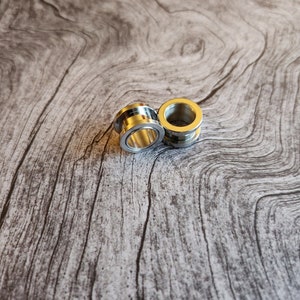 May include: Two silver metal ear gauges, one slightly larger than the other, on a woodgrain background.