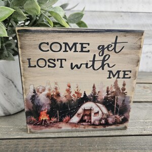 May include: Wooden sign with a watercolor illustration of a campsite with a tent, trees, and a campfire. The sign reads "Come get lost with me."