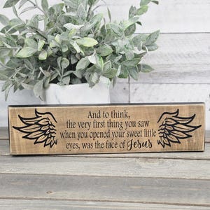 May include: Wooden sign with black text and angel wings. The text reads "And to think, the very first thing you saw when you opened your sweet little eyes, was the face of Jesus."