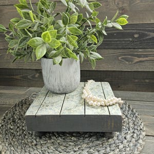 May include: A decorative wooden riser with a gray base and a patterned top, holding a potted plant with green leaves and a pearl necklace. The riser is on a woven mat, creating a rustic aesthetic.