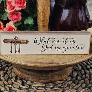 May include: A wooden sign with a white background and black lettering. The sign reads "Whatever it is, God is greater." There is a small image of a key rack with keys on it to the left of the text.