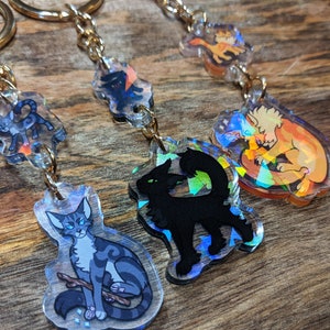 Può includere: Quattro portachiavi iridescenti con diversi personaggi di gatti. I portachiavi sono realizzati in acrilico trasparente con una finitura olografica. I personaggi includono un gatto grigio e bianco, un gatto nero, un gatto marrone e un gatto bianco con una coda marrone.