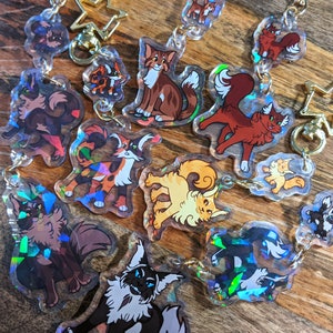 Może przedstawiać: Kolekcja breloków z holograficznymi, tęczowymi zawieszkami przedstawiającymi kreskówkowe koty. Zawieszki są w różnych pozach i kolorach, w tym czarnym, białym, pomarańczowym i brązowym.