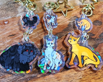 May include: Four iridescent keychains featuring cartoon cats. Each keychain has a gold star charm and a chain. The cats are a black cat, a white and orange cat, a white and gray cat, and a yellow cat.