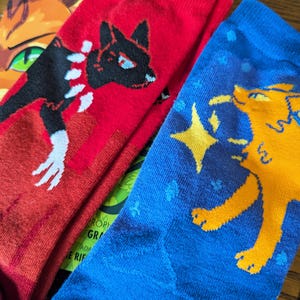 May include: Two pairs of socks with colorful graphic designs. One pair is red with a black cat design and the other pair is blue with a yellow cat design.