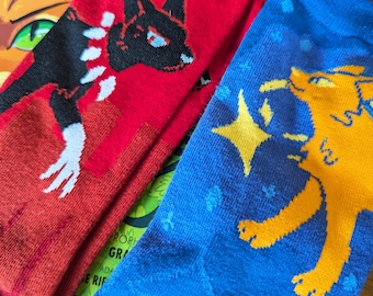 Può includere: Due paia di calzini con disegni grafici colorati. Un paio è rosso con un design di gatto nero e l'altro paio è blu con un design di gatto giallo.