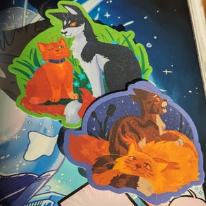 Op de afbeelding: Een verzameling kat-thema stickers. Eén sticker toont een oranje kat met een blauwe halsband en een zwart-witte kat. Een andere toont twee katten, één oranje en één bruin, tegen een nachtelijke hemel. De stickers hebben een glitterafwerking.