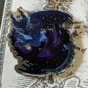 May include: A clear acrylic keychain featuring two stylized dragons. One dragon is dark blue with a starry wing, the other is black with purple accents. The dragons are intertwined, with a silver-colored clasp at the top.