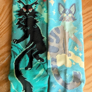 Könnte beinhalten: Zwei Socken mit Katzenmotiven. Eine Socke ist türkis mit einer schwarzen Katzenillustration, die andere hellblau mit einer grau-braunen Katzenillustration. Beide Socken haben einen detaillierten, cartoonartigen Stil.