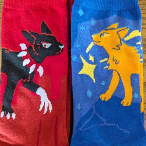 May include: A pair of socks with a black cat on one sock and an orange cat on the other sock. The cat on the red sock is facing left and the cat on the blue sock is facing right.