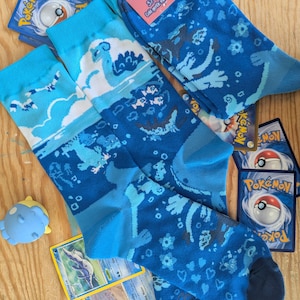 May include: Three pairs of blue socks with aquatic-themed designs, including waves, fish, and sea creatures. The socks are in shades of blue and white. Several Pokémon trading cards and a small blue toy are scattered around the socks.