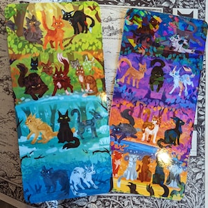 Op de afbeelding: Twee kleurrijke bladwijzers met illustraties van katten. Eén bladwijzer heeft een groene en blauwe achtergrond, de andere een paarse en oranje achtergrond. De katten zijn afgebeeld in verschillende kleuren, waaronder oranje, zwart, wit en grijs.