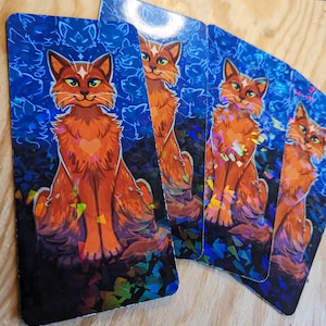 Pode incluir: Quatro cartas de tarot com uma ilustra&ccedil;&atilde;o de um gato laranja com um cora&ccedil;&atilde;o no peito. As cartas t&ecirc;m um fundo azul com silhuetas de gatos e detalhes hologr&aacute;ficos. As cartas est&atilde;o em leque sobre uma superf&iacute;cie de madeira.