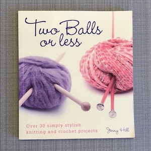 Op de afbeelding: Boekomslag met de titel "Two Balls or Less" in paarse tekst. De omslag toont twee bollen garen, een paarse en een roze, met breinaalden.