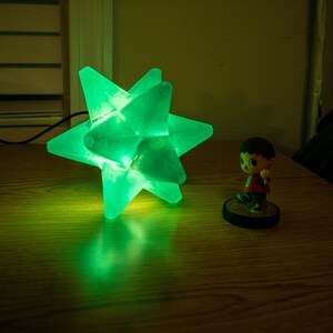 Op de afbeelding: Een groen oplichtende stervormige lamp staat op een houten oppervlak. Daarnaast een klein beeldje van een personage met een rood shirt en bruin haar. De lamp heeft een modern, geometrisch ontwerp. Afmetingen ca. 20 cm.