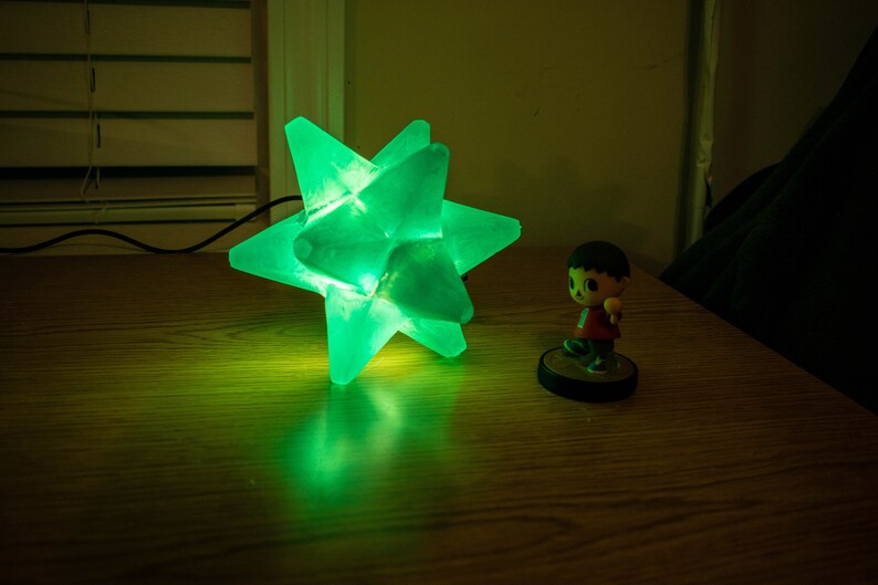 Op de afbeelding: Een groen oplichtende stervormige lamp staat op een houten oppervlak. Daarnaast een klein beeldje van een personage met een rood shirt en bruin haar. De lamp heeft een modern, geometrisch ontwerp. Afmetingen ca. 20 cm.