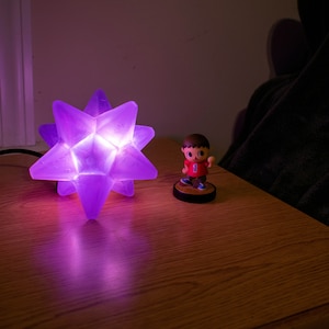 May include: A glowing purple star-shaped lamp sits on a wooden surface, casting a soft light. Beside it is a small figurine of a character in a red shirt. The lamp is plugged into a wall outlet. The scene is set in a dimly lit room.