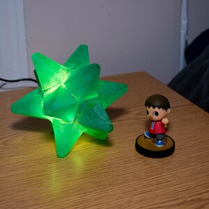 Op de afbeelding: Een groene, stervormige lamp met een gloeiend interieur staat op een houten oppervlak. Daarnaast staat een klein beeldje van een personage in een rood shirt en een blauwe korte broek. De lamp is aangesloten.