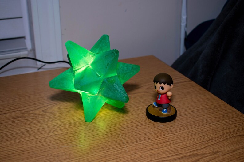 Op de afbeelding: Een groene, stervormige lamp met een gloeiend interieur staat op een houten oppervlak. Daarnaast staat een klein beeldje van een personage in een rood shirt en een blauwe korte broek. De lamp is aangesloten.