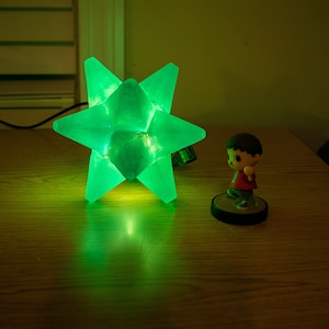 Op de afbeelding: Een heldergroene stervormige lamp op een houten oppervlak, die een groene gloed uitstraalt. Een klein beeldje van een personage in een rood shirt en bruin haar staat in de buurt. De lamp is aangesloten en verlicht.