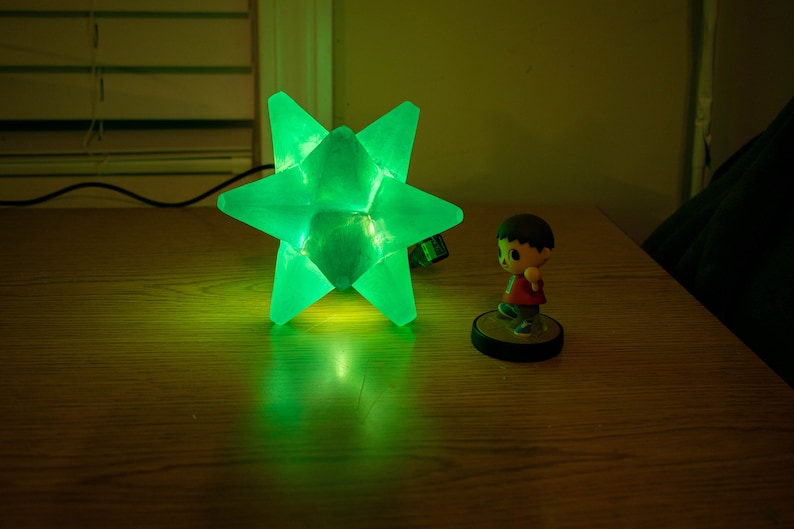 Op de afbeelding: Een heldergroene stervormige lamp op een houten oppervlak, die een groene gloed uitstraalt. Een klein beeldje van een personage in een rood shirt en bruin haar staat in de buurt. De lamp is aangesloten en verlicht.
