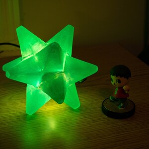 Op de afbeelding: Een heldergroene stervormige lamp staat op een houten oppervlak. Daarnaast staat een klein beeldje van een stripfiguur. Het geometrische ontwerp en de heldere kleur van de lamp cre&euml;ren een opvallend contrast.