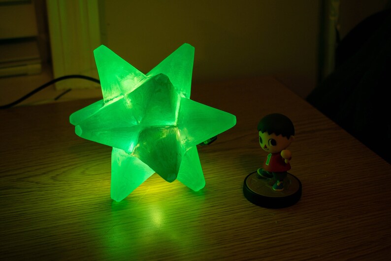 Op de afbeelding: Een heldergroene stervormige lamp staat op een houten oppervlak. Daarnaast staat een klein beeldje van een stripfiguur. Het geometrische ontwerp en de heldere kleur van de lamp cre&euml;ren een opvallend contrast.