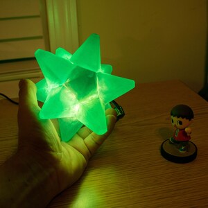 Op de afbeelding: Een gloeiende groene stervormige lamp, vastgehouden in een hand. De lamp is gemaakt van doorschijnend materiaal en straalt een helder groen licht uit. Een klein beeldje van een personage in een rood shirt en bruin haar staat op een ronde basis.