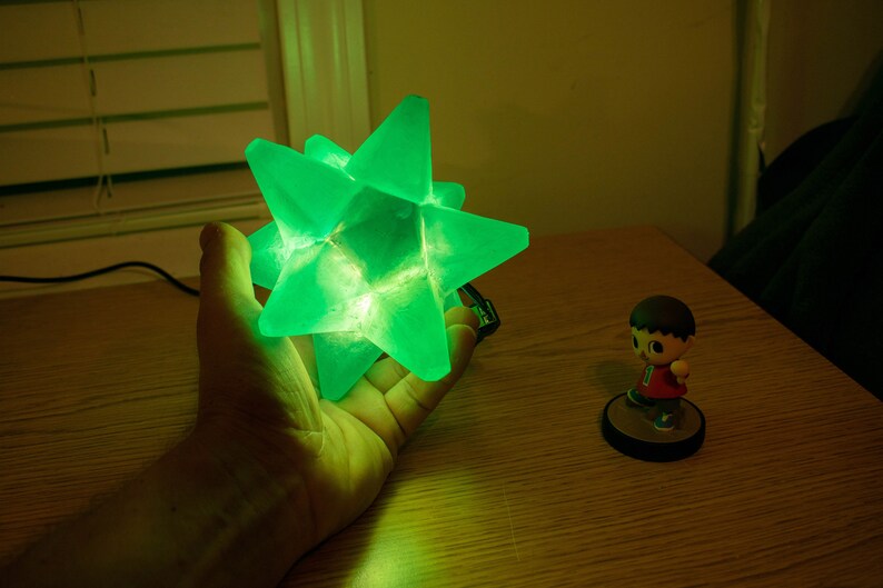 Op de afbeelding: Een gloeiende groene stervormige lamp, vastgehouden in een hand. De lamp is gemaakt van doorschijnend materiaal en straalt een helder groen licht uit. Een klein beeldje van een personage in een rood shirt en bruin haar staat op een ronde basis.