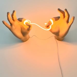 May include: Sculptural wall art featuring two hand-shaped forms in a neutral tone, seemingly holding a glowing, warm-toned light. The light creates a visual connection between the hands, with a wire extending downwards.