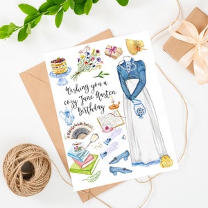 May include: A white card with a watercolor illustration of a woman in a blue dress and white skirt. The illustration is surrounded by various objects, including a book, a teacup, a fan, and a bouquet of flowers. The text "Wishing you a cozy Jane Austen birthday" is written in black cursive.