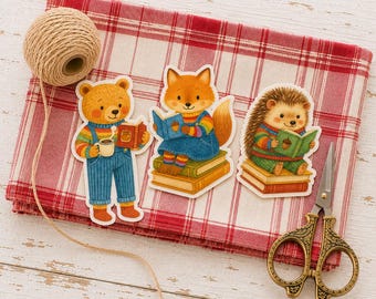 Op de afbeelding: Drie gestanste stickers met een bruine beer met een mok, een vos die een boek leest en een egel die een boek leest, allemaal met kleurrijke truien. De stickers liggen op een rood-wit geruite doek met een bolletje touw en een schaar.