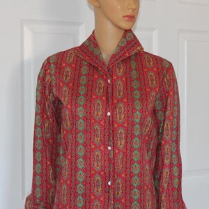 May include: A red long-sleeve button-down shirt with a repeating pattern of green, yellow, and purple shapes. The shirt has a wide collar and white buttons. The sleeves are gathered at the cuffs.