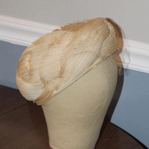 May include: A vintage beige hat with a net veil, displayed on a mannequin head. The hat features a textured design and a delicate, sheer veil. The hat is a classic accessory, likely from a bygone era.