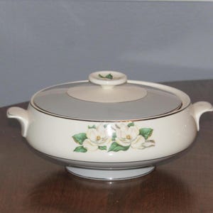 May include: A white ceramic serving bowl with a lid, featuring a floral design of white magnolia blossoms and green leaves. The bowl has a light blue interior and a gold trim.
