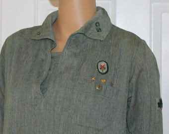 May include: A green long-sleeved shirt with a notched collar and a small pocket. The shirt features a patch with a flower design, small pins, and the letters "C.S." on the collar. A green shamrock patch is on the sleeve.