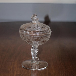 May include: A clear glass candy dish with a lid and a decorative swirl design. The dish has a pedestal base with a leaf-shaped design.