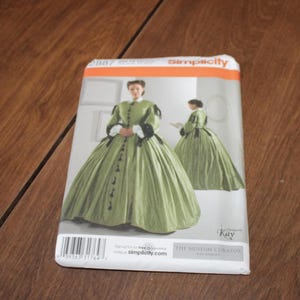 May include: Simplicity sewing pattern featuring a green historical dress design. The pattern cover shows a woman in a long, flowing dress with black accents. The pattern is from The Museum Curator collection.