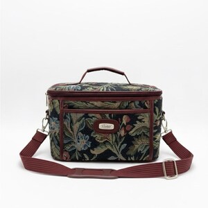 May include: A floral tapestry cosmetic bag with a burgundy handle and trim. The bag has a detachable burgundy shoulder strap and silver hardware. The bag features a dark background with a floral pattern in shades of green, blue, and tan.