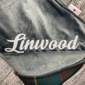 May include: A gray plush blanket with the name "Linwood" embroidered in white script. The blanket has a plaid pattern on the underside with green, red, and yellow accents. A small tag with a red maple leaf is visible.
