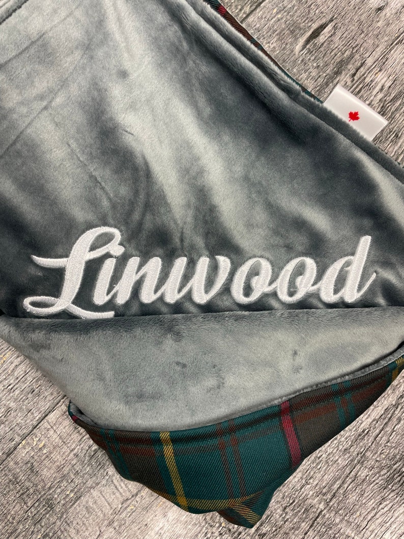 May include: A gray plush blanket with the name "Linwood" embroidered in white script. The blanket has a plaid pattern on the underside with green, red, and yellow accents. A small tag with a red maple leaf is visible.