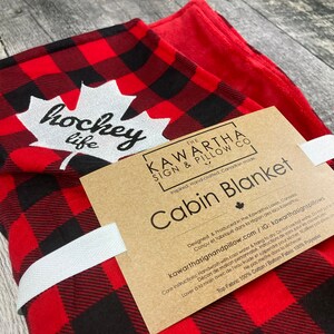 May include: Red and black checked cabin blanket with a white maple leaf and the text "hockey life". The blanket is made in Canada and is hand-crafted. The tag says "The Kawartha Sign & Pillow Co."
