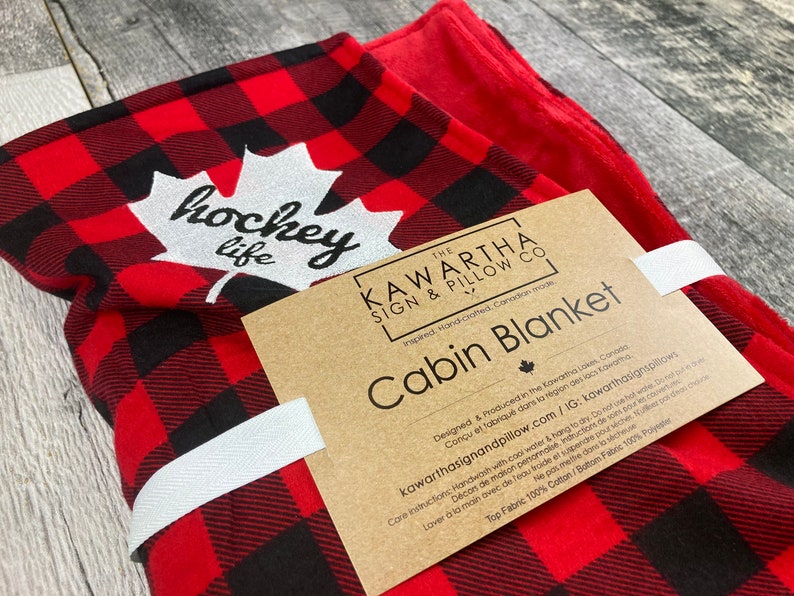 May include: Red and black checked cabin blanket with a white maple leaf and the text "hockey life". The blanket is made in Canada and is hand-crafted. The tag says "The Kawartha Sign & Pillow Co."
