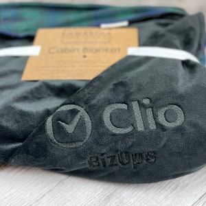 May include: A dark gray plush cabin blanket with the embroidered word "Clio" and the BizOps logo. The blanket is folded and tied with a white ribbon, with a brown tag that reads "Cabin Blanket". A folded plaid blanket is visible in the background.