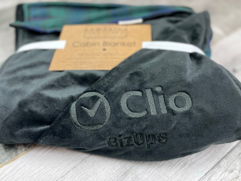 May include: A dark gray plush cabin blanket with the embroidered word "Clio" and the BizOps logo. The blanket is folded and tied with a white ribbon, with a brown tag that reads "Cabin Blanket". A folded plaid blanket is visible in the background.