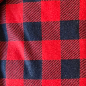 May include: Close-up of a red and black buffalo plaid fabric. The pattern consists of large, evenly spaced squares in a classic check design. The fabric has a soft, textured appearance.