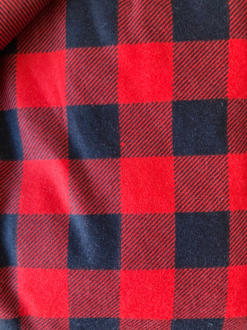 May include: Close-up of a red and black buffalo plaid fabric. The pattern consists of large, evenly spaced squares in a classic check design. The fabric has a soft, textured appearance.