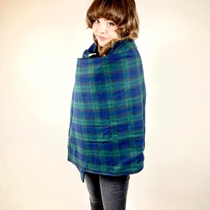 May include: A blue and green plaid cape with a button closure. The cape is wrapped around a person. The person is wearing black jeans. The cape has a classic plaid pattern in shades of blue and green.