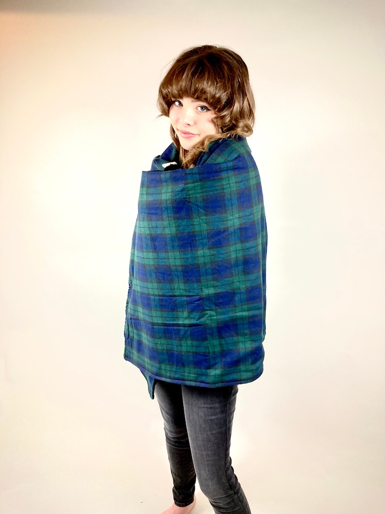 May include: A blue and green plaid cape with a button closure. The cape is wrapped around a person. The person is wearing black jeans. The cape has a classic plaid pattern in shades of blue and green.